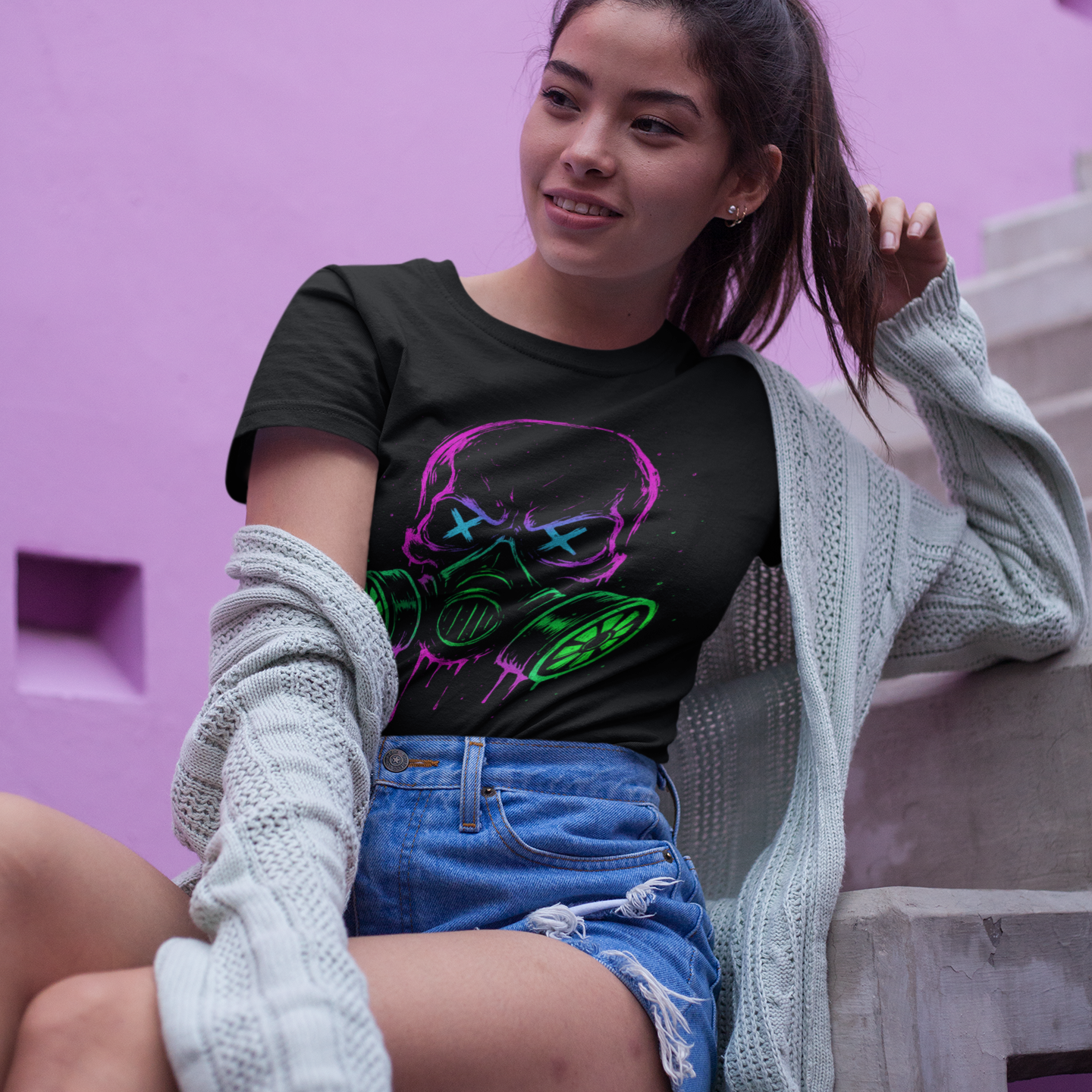 Woman wearing a black t-shirt with colorful design and denim shorts, sitting against a pink wall.