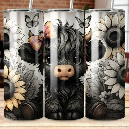 Tumbler with a design of a black cow with a bow, surrounded by flowers and butterflies on a wooden surface.
