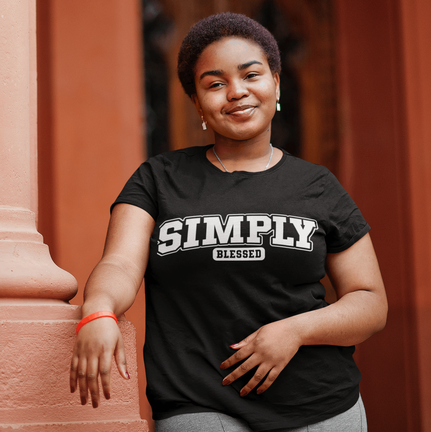 Woman wearing a black t-shirt with 'Simply Blessed' text, standing against a red brick wall.