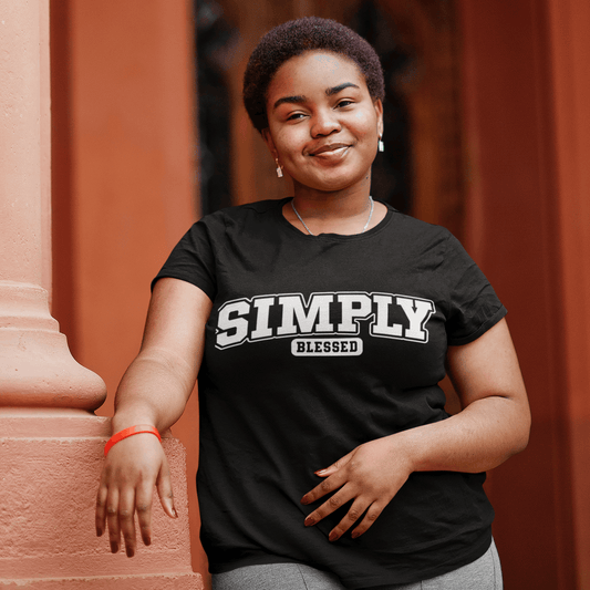 Woman wearing a black t-shirt with 'Simply Blessed' text, standing against a red brick wall.