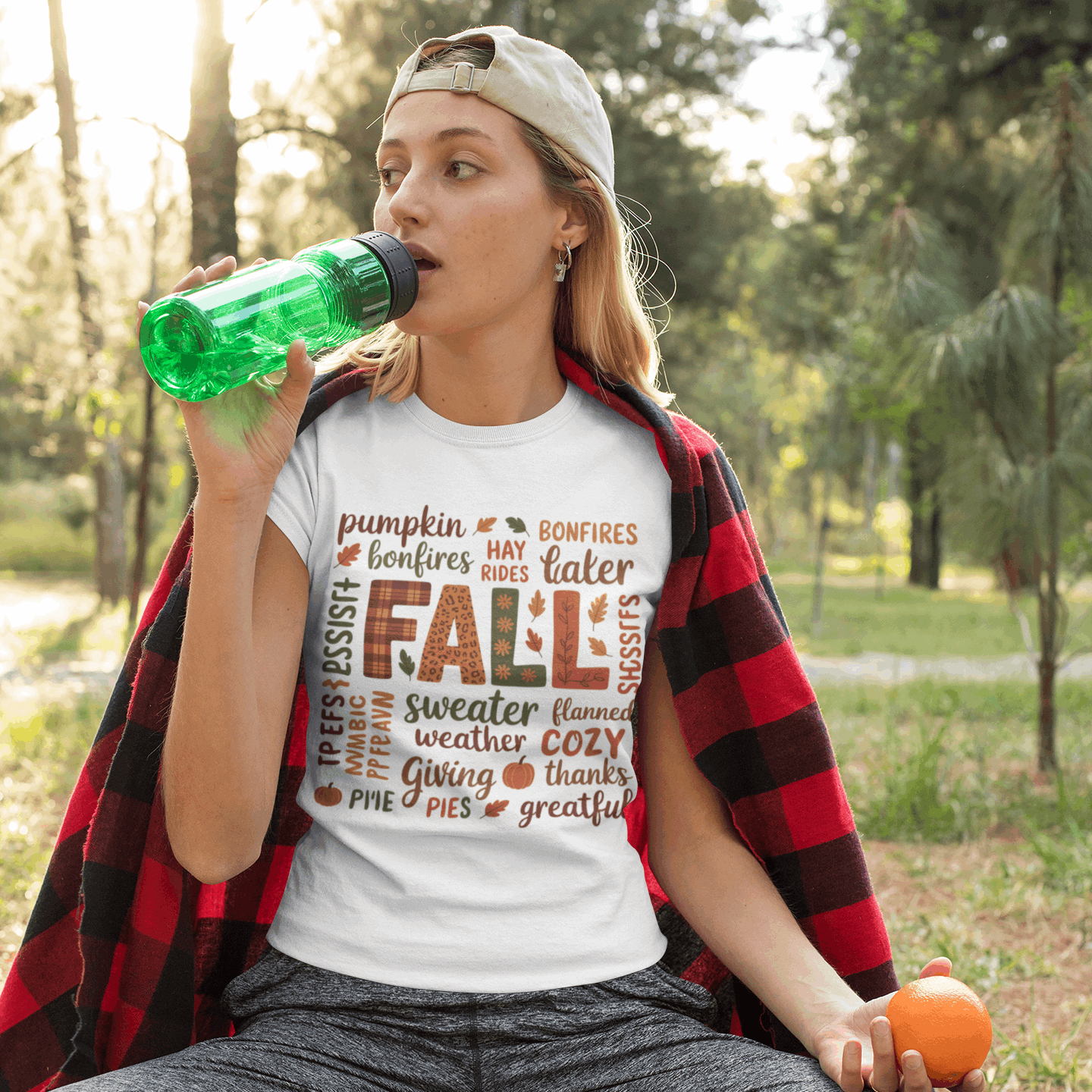Woman sitting outdoors wearing a 'Fall' themed t-shirt, holding an orange and a green bottle.