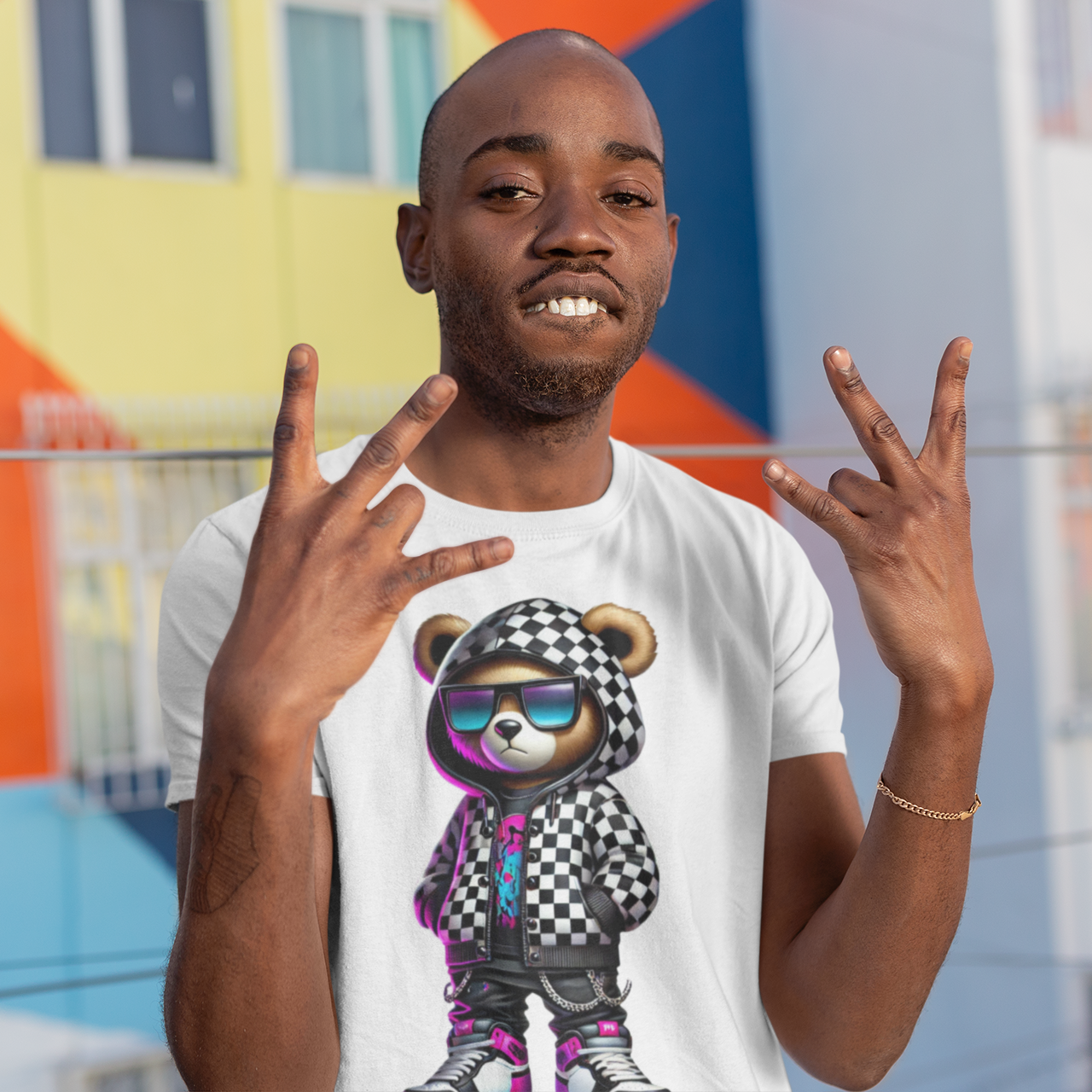 Man wearing a white t-shirt with a graphic design of a bear in sunglasses and a checkered hat, making peace signs in front of a colorful mural.