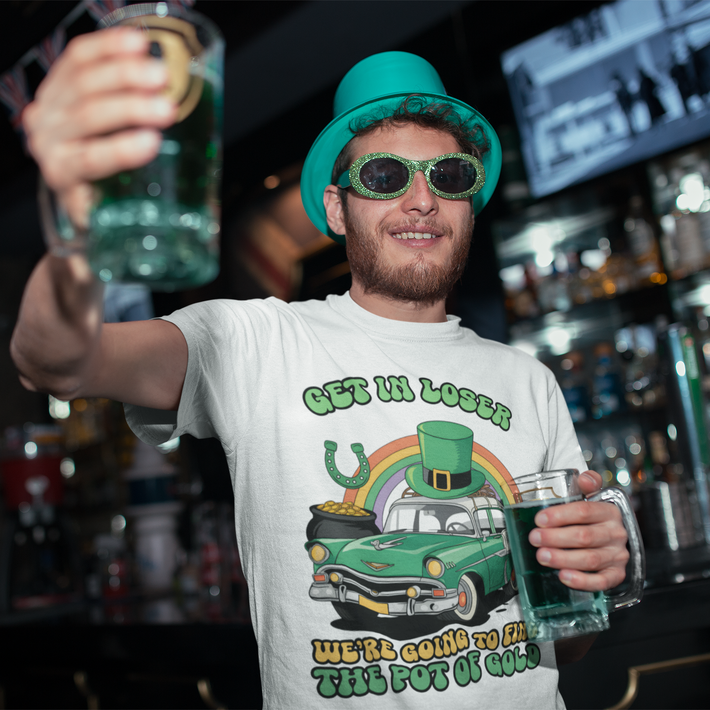 Man in a bar wearing a green hat and sunglasses, holding two green beers, with a shirt featuring a car and pot of gold graphic.