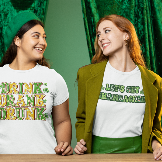 Two women wearing white t-shirts with humorous text designs, one holding a beer, against a green curtain background.