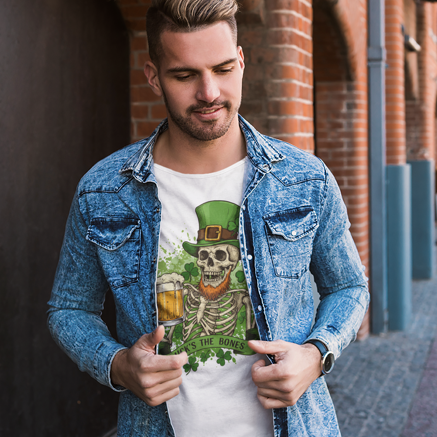 Man wearing a denim jacket over a white t-shirt with a graphic design, standing against a brick wall.