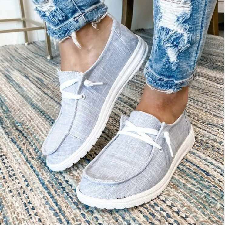 Gray slip-on shoes with white laces worn with ripped jeans on a textured floor.